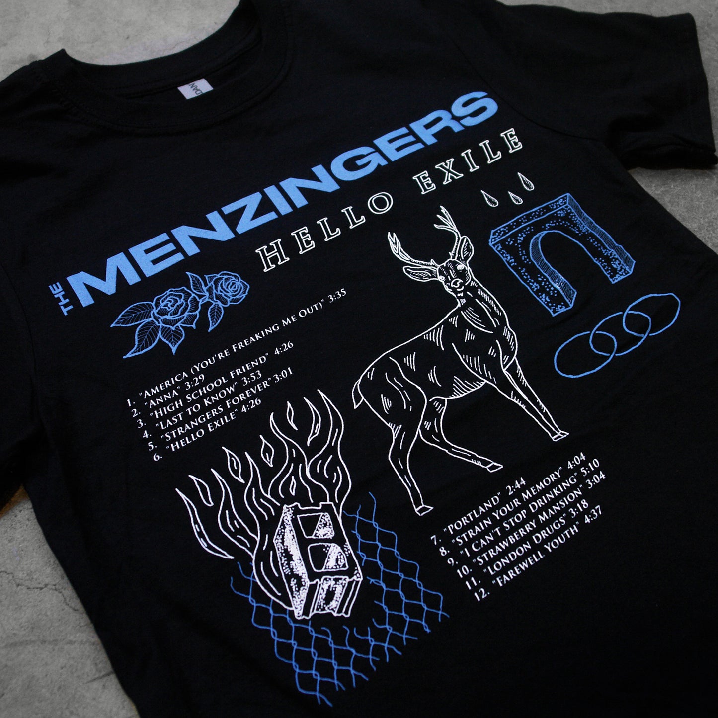 angled close up image of black tee shirt laid flat on cement floor as background. tee has full chest print. along the top in blue saus the menzingers and has several images below. including blue flower, white deer, the words hello exile in white, and cinder block and the track list to the menzingers album hello exile.