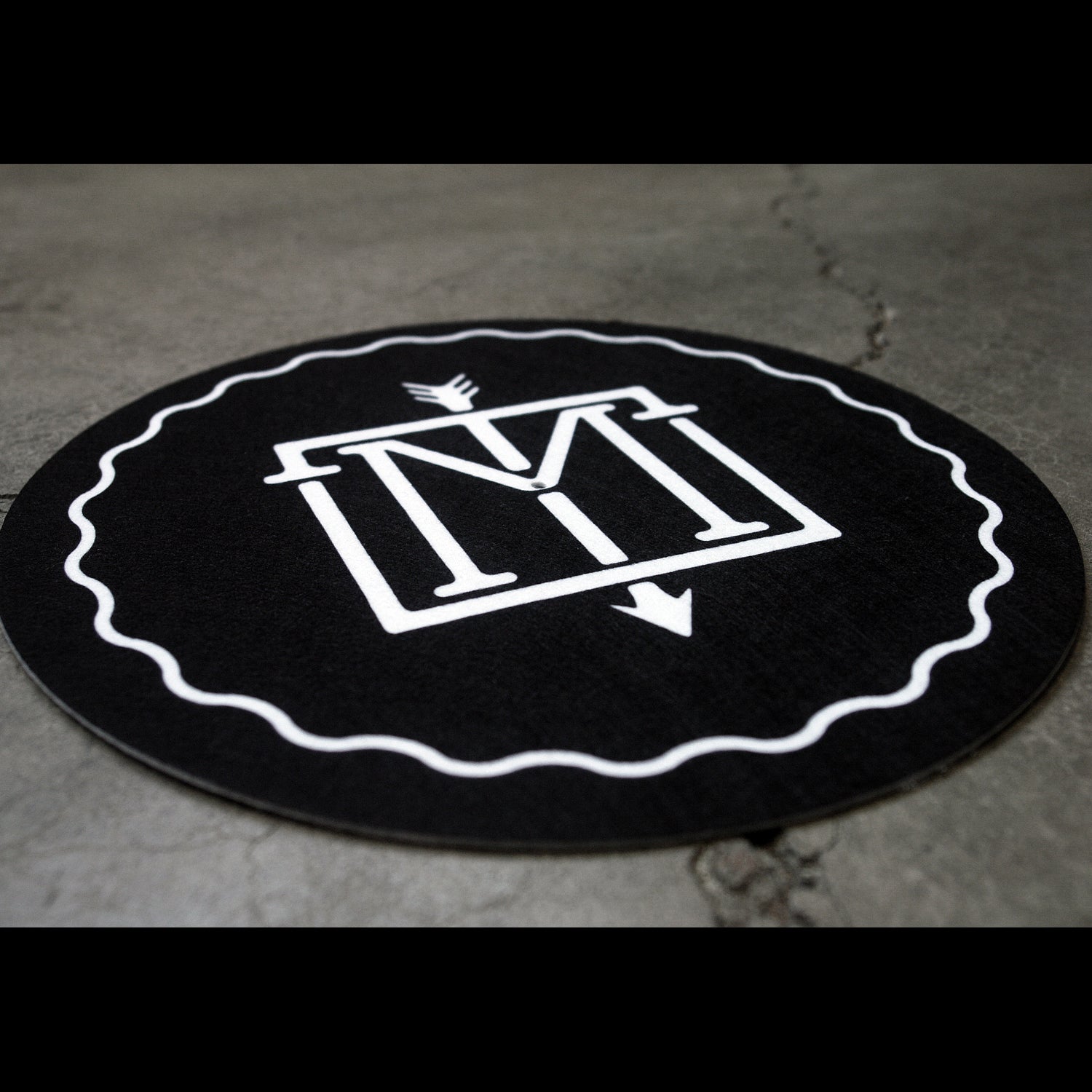 angled close up image of black slipmat for record player laid flat on cement floor. slipmat is a circle with white swirl design framing the outside and the center has an emblem with the letter M and an arrow down the center.