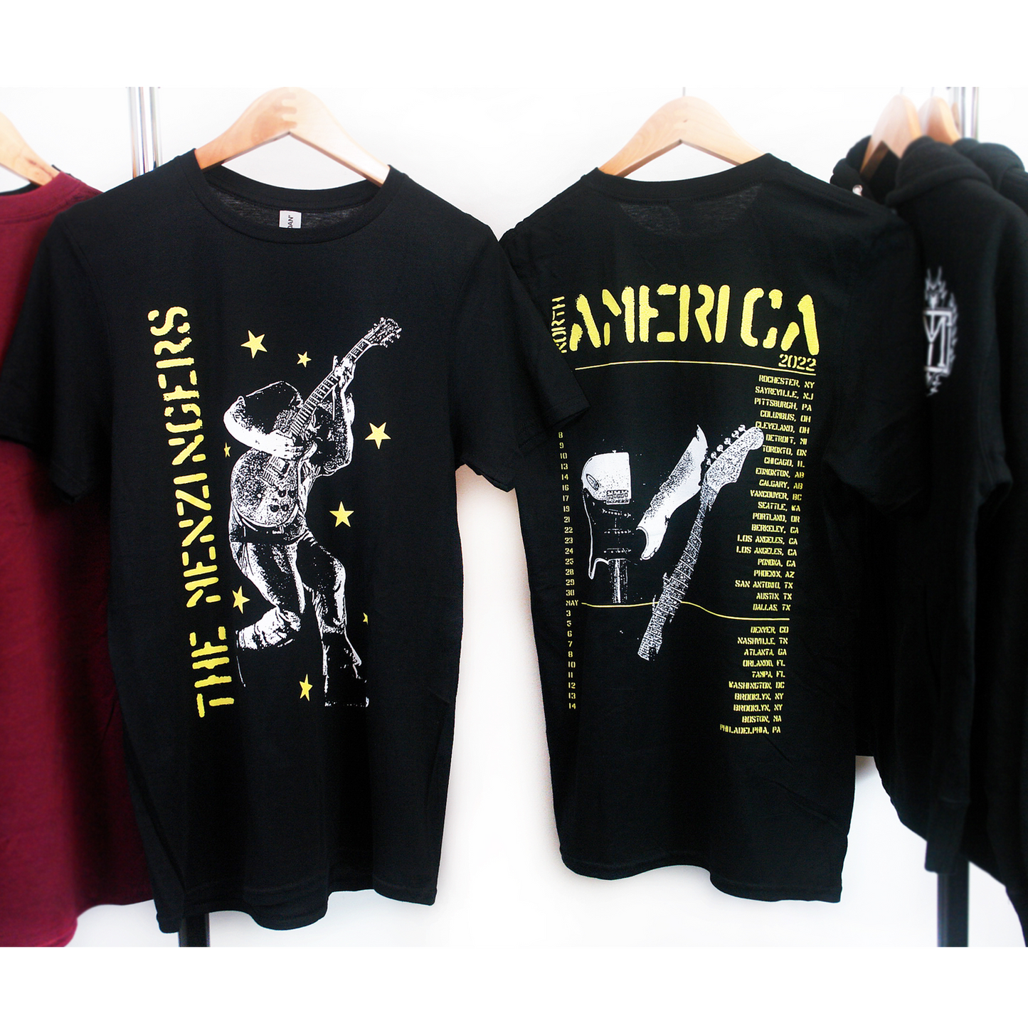 image of the front and back of a black tee shirt hanging on a white background. front of tee is on the left and has a full body print in yellow on the left side vertically says the menzingers with yellow stars all over and in white an image of a man playing a guitar. the back of the tee is on the right and has in yellow at the top 2022 america with the list of tour dates and locations below and a broken guitar in the center