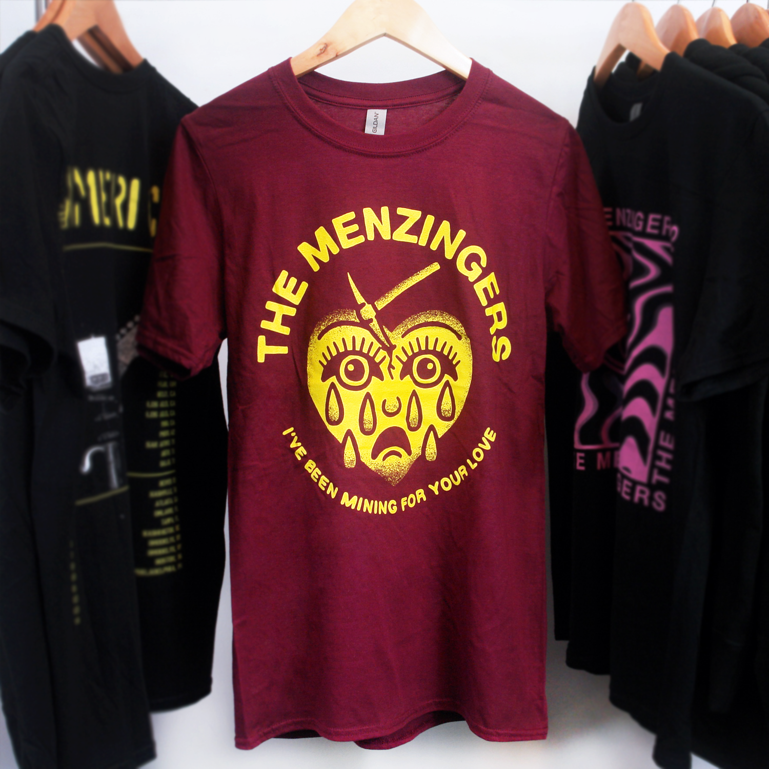 image of a maroon tee shirt hanging on a white background. the tee shirt has a full center chest print in yellow of a heart with a sad face and tears and a mining pick in the top. arhced around the heart says the menzingers. arhced around at the bottom of the heart says i've been mining for your love